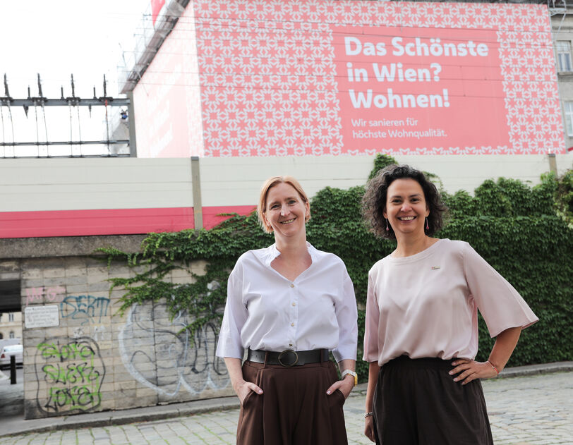 Zwei Frauen stehen vor einer mit Graffiti besprühten Mauer und einem Plakat mit der Aufschrift 'Das Schönste in Wien? Wohnen!'