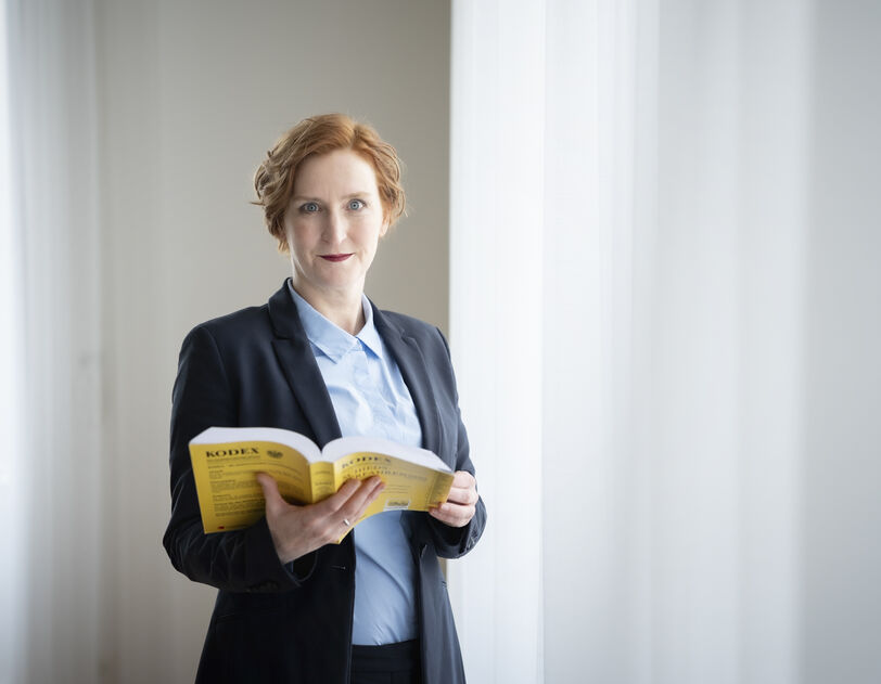 Frau mit roten Haaren in dunklem Blazer und hellblauer Bluse steht vor hellem Vorhang und hält ein aufgeschlagenes gelbes Buch, den Mietrechts-Kodex, in den Händen