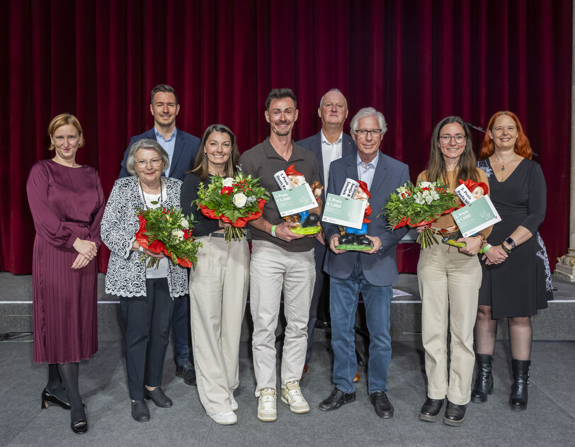 Gruppe von acht Personen vor rotem Vorhang und österreichischer Flagge, drei Personen halten Blumensträuße und Urkunden